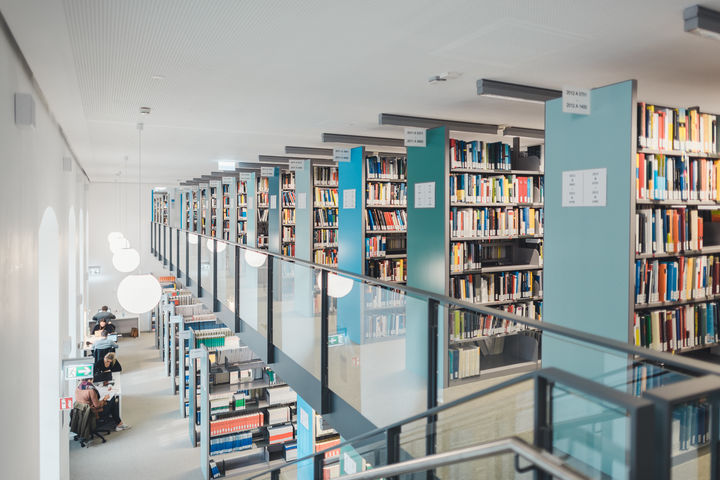 Blick auf die Galerie mit Bücherregalen und auf die Lehrbuchsammlung im Erdgeschoss im Ausleihzentrum Schloss Westflügel. Die Regalblenden sind in Blau- und Türkistönen gehalten, an der Decke hängen runde weiße Lampen.
