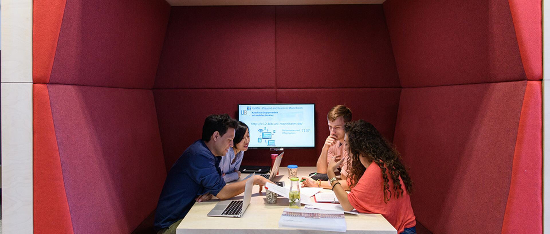 Vier Studierende sitzen in einem roten Kubus im Learning Center im Bibliotheksbereich Schloss Schneckenhof. An der Rückwand des Kubus hängt ein PalMA-Team-Monitor.