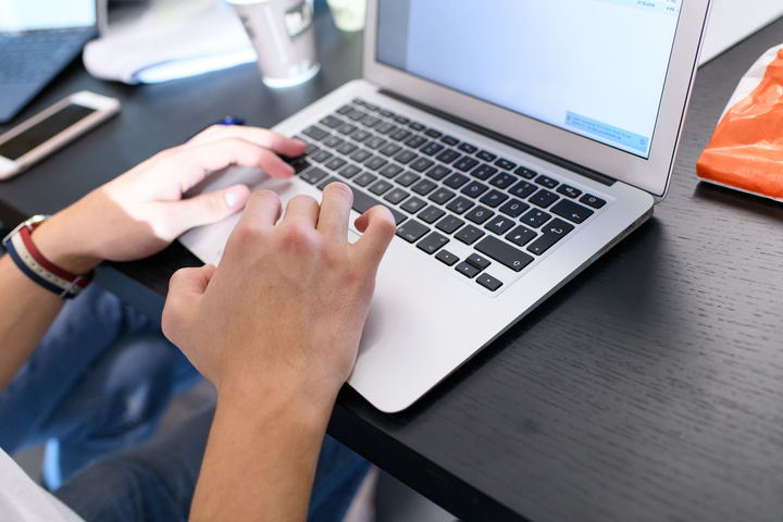 Eine Person, deren Hände im Bild sind, schreibt an einem Laptop. Neben dem Laptop sind ein Smartphone und ein Kaffeebecher auf dem Tisch.
