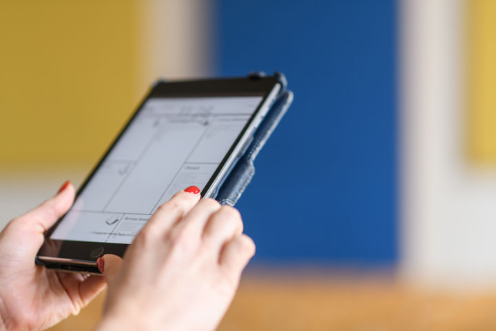 Ein E-Book-Reader wird in zwei Händen gehalten. Im Hintergrund sind gelbe und blaue Farbelemente an der Wand.