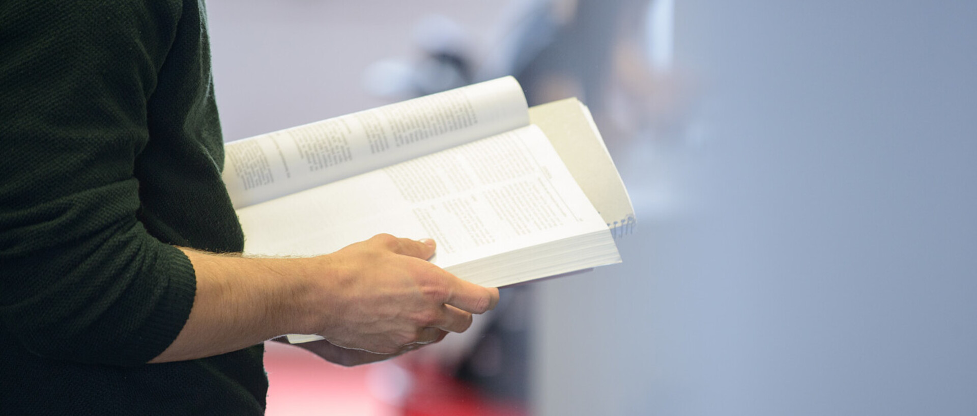 Eine Person steht in der Bibliothek und hält ein aufgeschlagenes Buch in den Händen.