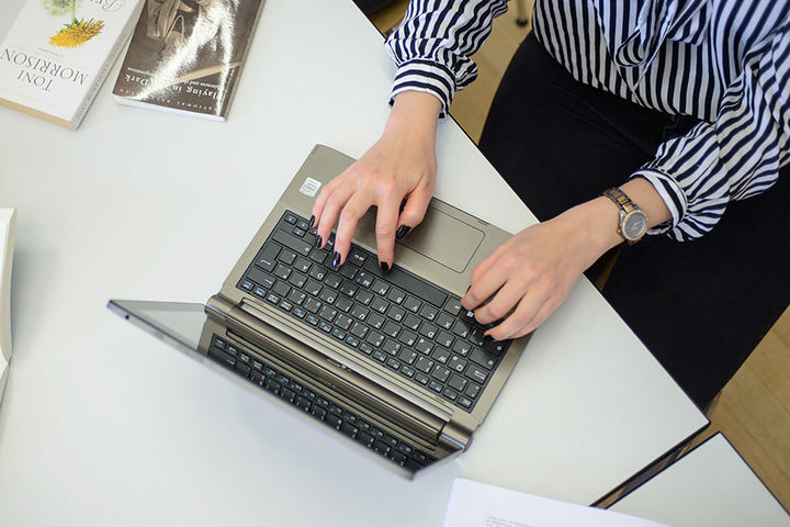 Eine Frau schreibt an einem Laptop. Sie trägt eine schwarz-weiß gemusterte Bluse, eine schwarze Hose und am rechten Handgelenk eine Uhr. Ihre Fingernägel sind schwarz lackiert. Neben dem Laptop liegen ein aufgeschlagenes Buch, auf dem ein Stift und eine Brille abgelegt sind, weitere Bücher und ein schwarzes Schlüsselband mit weißem Aufdruck "Universität Mannheim" und dem Logo.